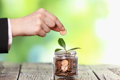 Photo of Investment concept. Woman putting coin into glass jar at wooden table against blurred green background, closeup