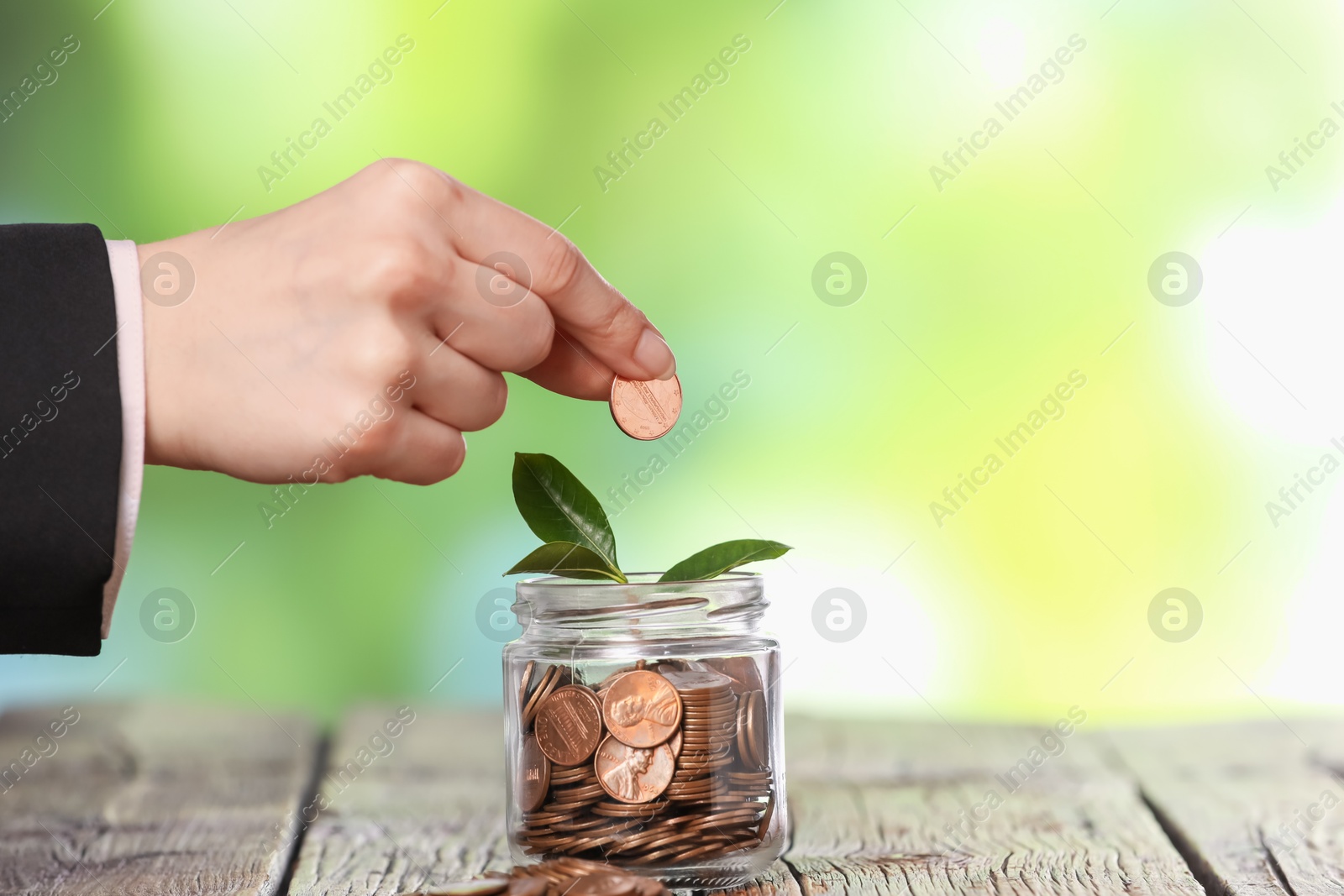 Investment concept. Woman putting coin into glass jar at wooden table against blurred green background, closeup Photo of Investment concept. Woman putting coin into glass jar at wooden table against blurred green background, closeup