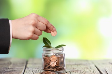 Photo of Investment concept. Woman putting coin into glass jar at wooden table against blurred green background, closeup