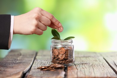 Photo of Investment concept. Woman putting coin into glass jar at wooden table against blurred green background, closeup