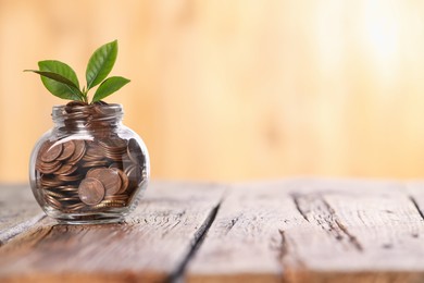 Photo of Investment concept. Glass jar with coins and sprout on wooden table against blurred orange background, space for text