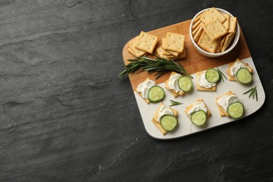 Photo of Tasty crackers with cream cheese, rosemary and cucumber on dark textured table, top view. Space for text