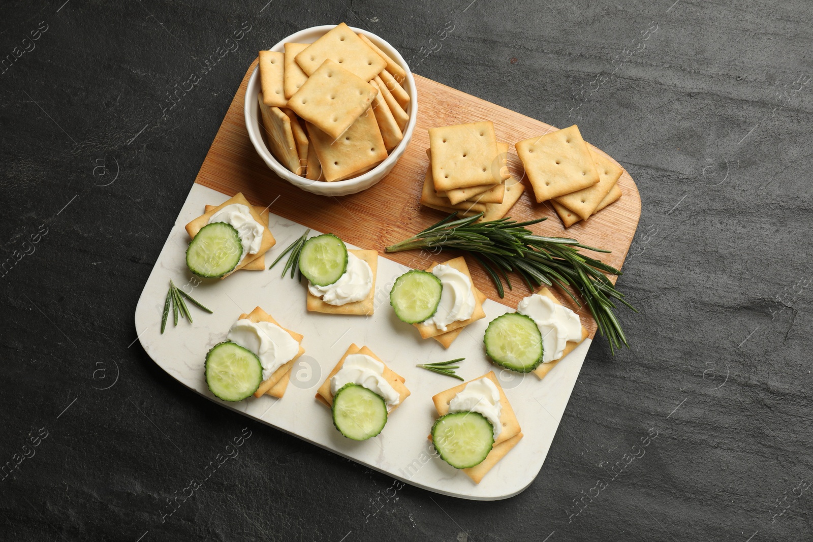 Photo of Tasty crackers with cream cheese, rosemary and cucumber on dark textured table, top view