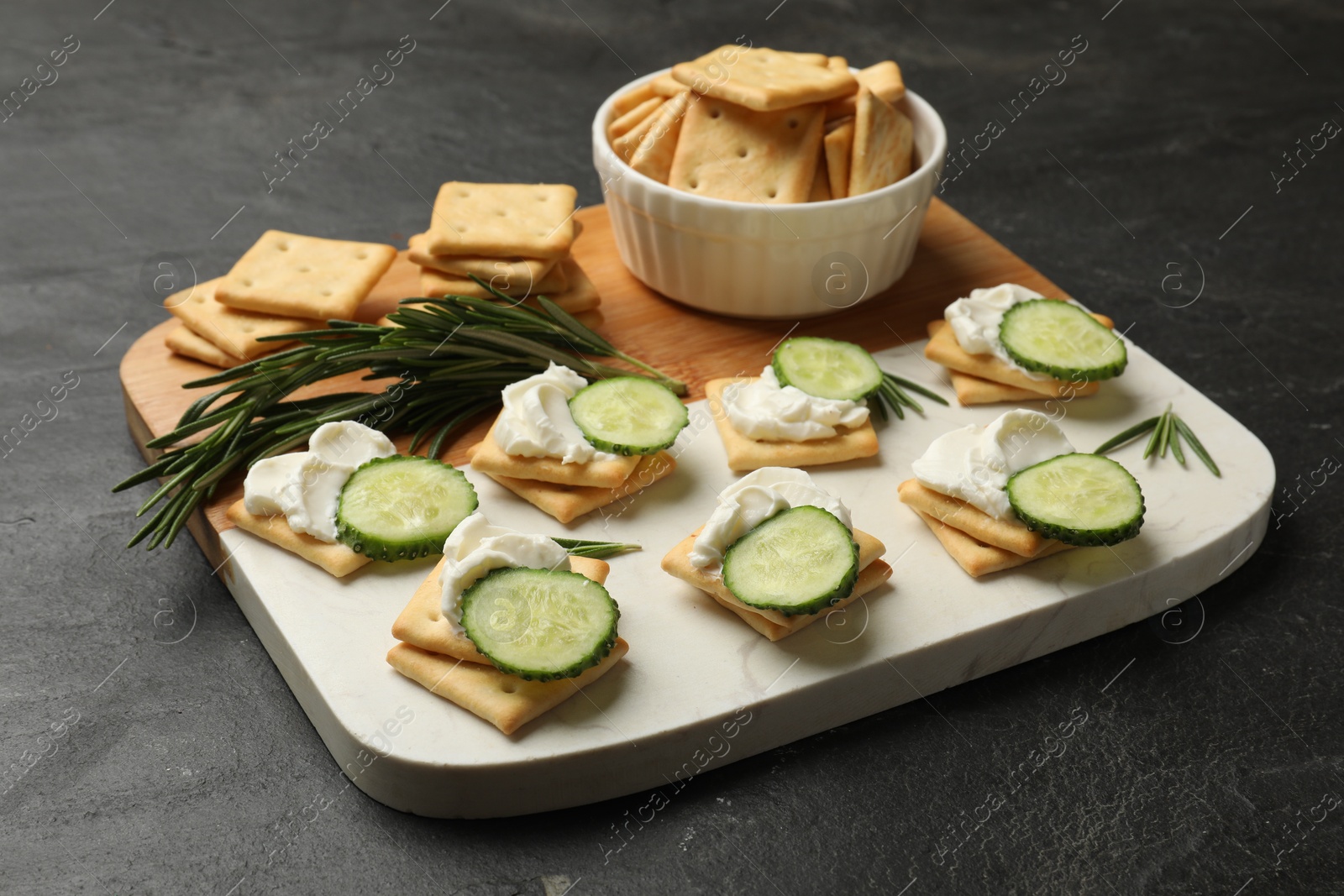 Photo of Tasty crackers with cream cheese, rosemary and cucumber on dark textured table, closeup