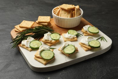 Photo of Tasty crackers with cream cheese, rosemary and cucumber on dark textured table, closeup