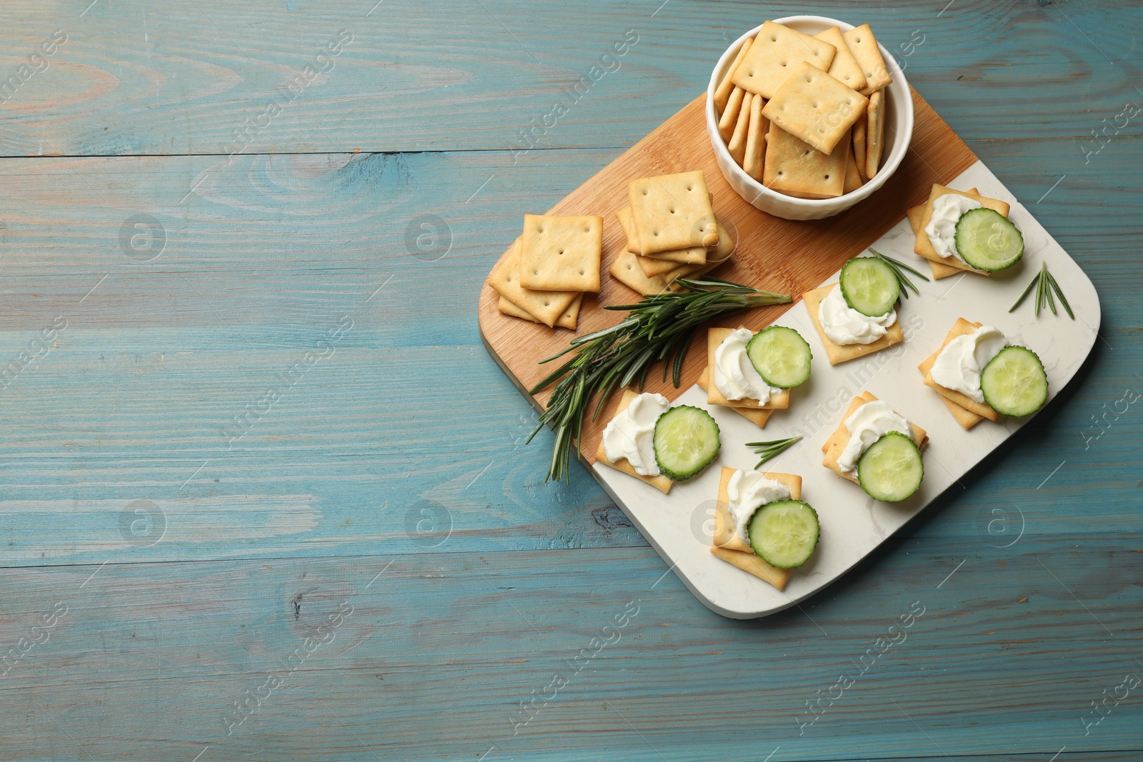 Photo of Tasty crackers with cream cheese, rosemary and cucumber on light blue wooden table, top view. Space for text