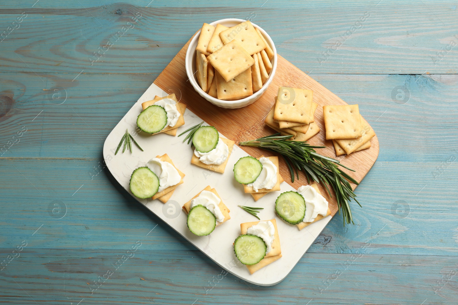 Photo of Tasty crackers with cream cheese, rosemary and cucumber on light blue wooden table, top view