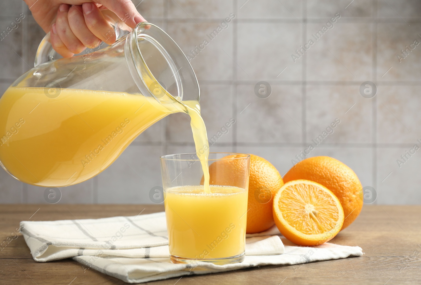 Woman pouring fresh orange juice from jug into glass at wooden table against grey tiled wall, closeup Photo of Woman pouring fresh orange juice from jug into glass at wooden table against grey tiled wall, closeup