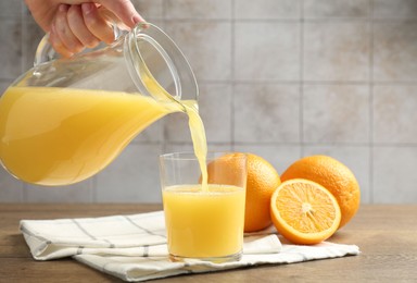 Photo of Woman pouring fresh orange juice from jug into glass at wooden table against grey tiled wall, closeup