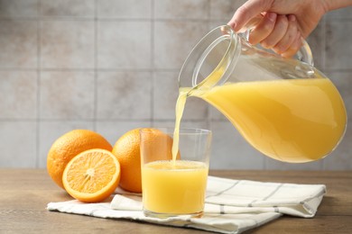 Photo of Woman pouring fresh orange juice from jug into glass at wooden table against grey tiled wall, closeup