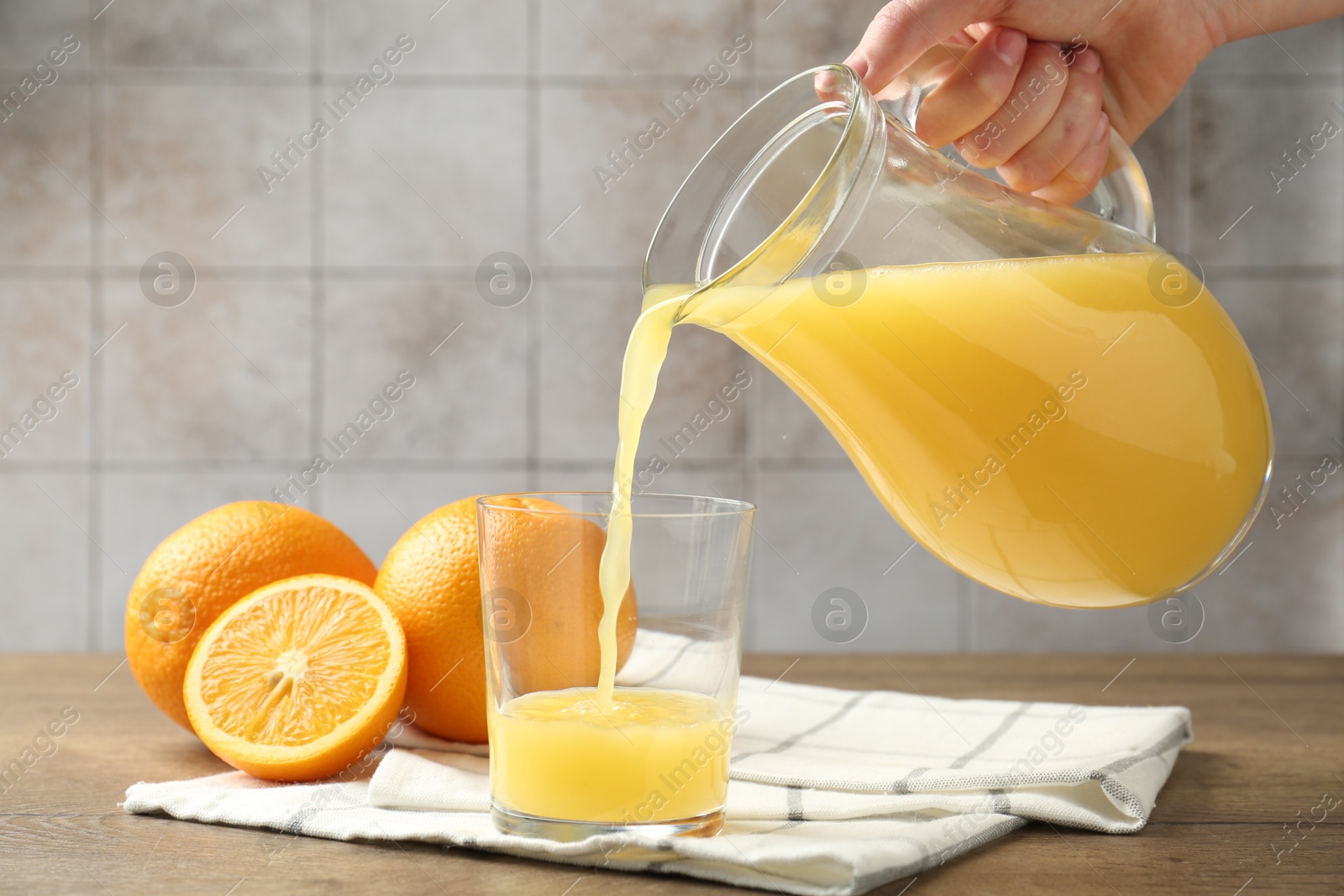 Woman pouring fresh orange juice from jug into glass at wooden table against grey tiled wall, closeup Photo of Woman pouring fresh orange juice from jug into glass at wooden table against grey tiled wall, closeup
