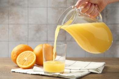 Photo of Woman pouring fresh orange juice from jug into glass at wooden table against grey tiled wall, closeup