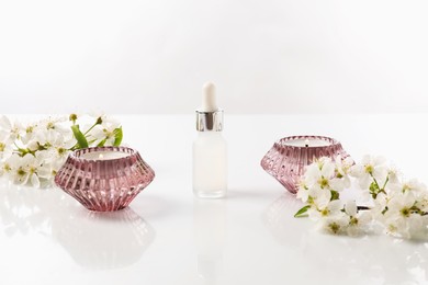 Photo of Bottle of cosmetic product, burning candles and beautiful flowers on white background, closeup