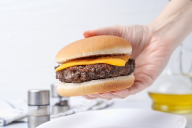 Photo of Woman holding tasty cheeseburger at white table, closeup