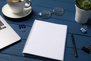 Photo of Notebook with pen, laptop, glasses, paper clips and coffee on blue wooden table, closeup