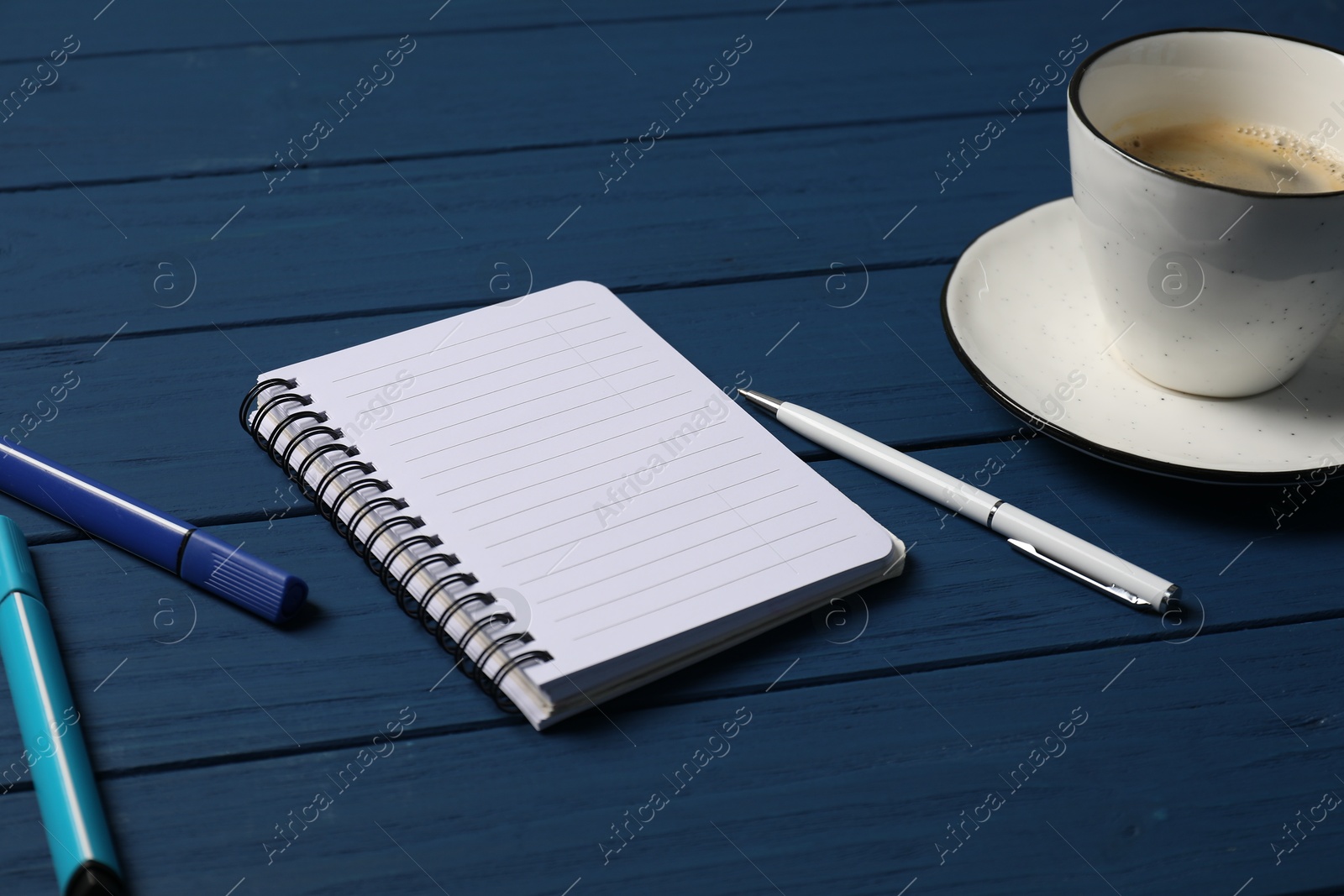 Photo of Notebook with pen, markers and coffee on blue wooden table, closeup
