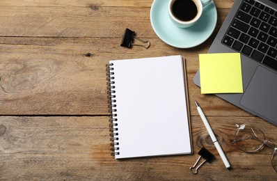Photo of Notebook with pen, sticky note, laptop, paper clips and coffee on wooden table, flat lay. Space for text