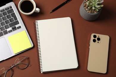 Photo of Notebook with pen, laptop, smartphone, sticky note and coffee on brown background, flat lay