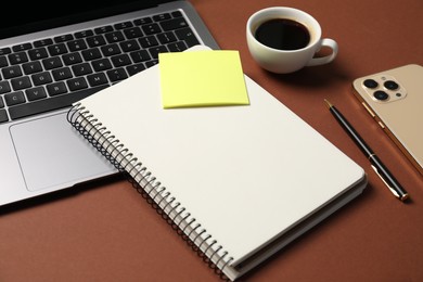 Photo of Notebook with pen, laptop, smartphone, sticky note and coffee on brown background, closeup