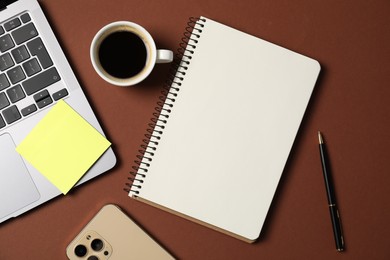 Photo of Notebook with pen, laptop, smartphone, sticky note and coffee on brown background, flat lay