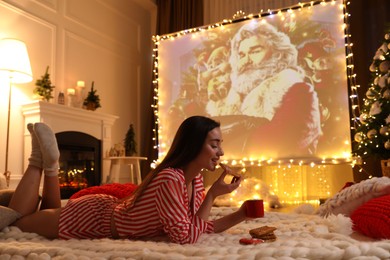 Photo of MYKOLAIV, UKRAINE - DECEMBER 24, 2020: Woman watching The Christmas Chronicles movie via video projector in room. Cozy winter holidays atmosphere