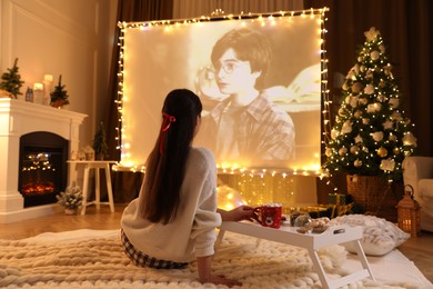 Photo of MYKOLAIV, UKRAINE - DECEMBER 24, 2020: Woman watching Harry Potter and Philosopher's Stone movie via video projector in room. Cozy winter holidays atmosphere