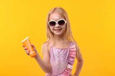 Photo of Smiling little girl with tube of sun protection cream against yellow background