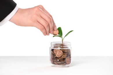 Photo of Investment concept. Woman putting coin into glass jar at white table against light background, closeup