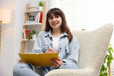 Photo of Woman writing in planner on armchair indoors