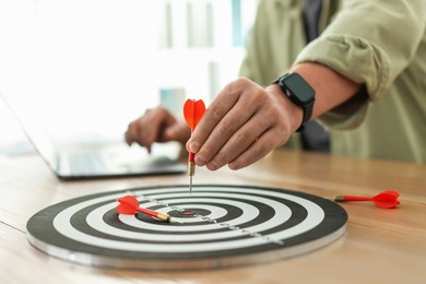 Photo of Man with dart aiming at dartboard while working on laptop indoors, closeup
