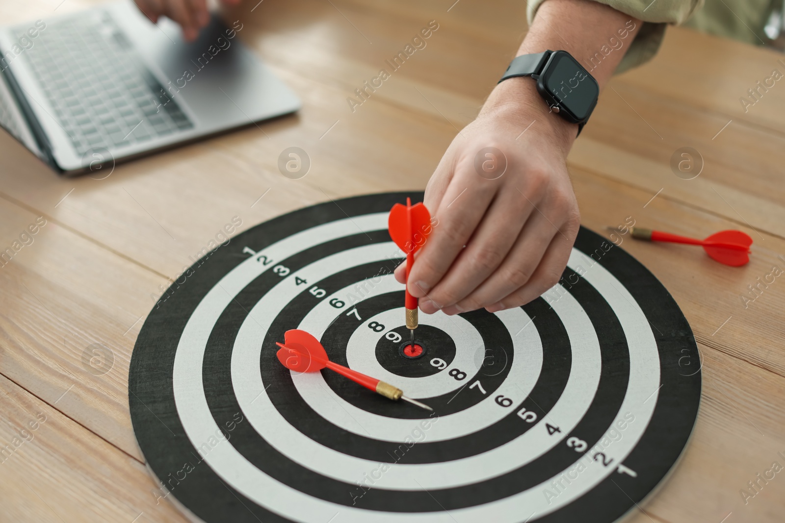 Photo of Man with dart aiming at dartboard while working on laptop indoors, closeup