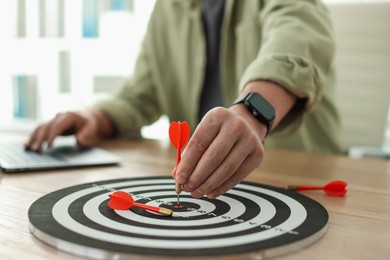 Photo of Man with dart aiming at dartboard while working on laptop indoors, closeup
