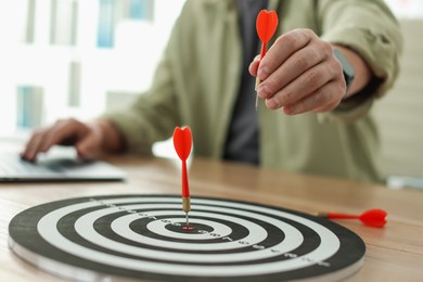 Photo of Man with dart aiming at dartboard while working on laptop indoors, closeup