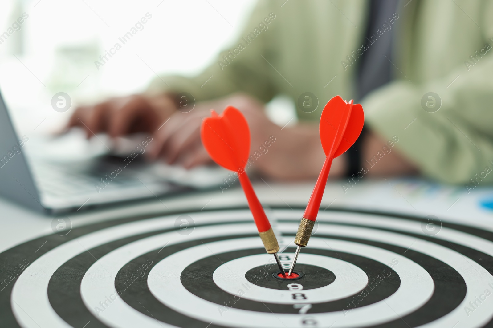 Photo of Man working on laptop at table, focus on dartboard with arrows indoors