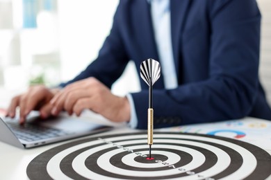 Photo of Man working on laptop at table, focus on dartboard with arrow indoors
