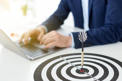 Photo of Man working on laptop at table, focus on dartboard with arrow indoors