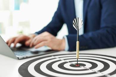Photo of Man working on laptop at table, focus on dartboard with arrow indoors