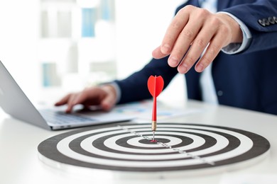 Photo of Man working on laptop at table, focus on dartboard with arrow indoors