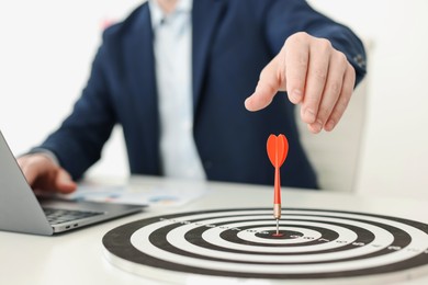 Photo of Man working on laptop at table, focus on dartboard with arrow indoors