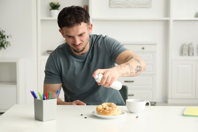 Photo of Emotional young man spraying insecticide onto pretzel with cockroaches at white table indoors. Pest problem