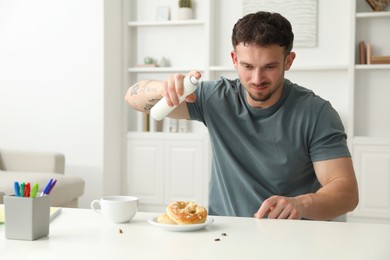 Photo of Emotional young man spraying insecticide onto pretzel with cockroaches at white table indoors. Pest problem