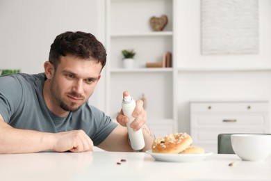 Photo of Emotional young man spraying insecticide onto pretzel with cockroaches at white table indoors. Pest problem