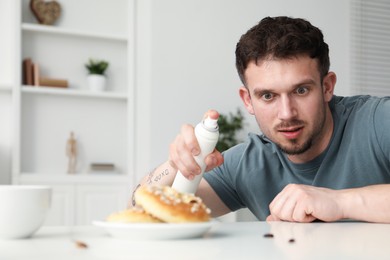 Photo of Emotional young man spraying insecticide onto pretzel with cockroaches at white table indoors. Pest problem
