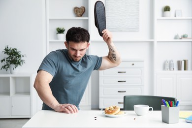 Photo of Emotional young man killing cockroaches with slipper at white table indoors. Pest problem