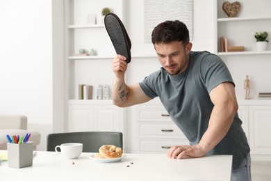 Photo of Emotional young man killing cockroaches with slipper at white table indoors. Pest problem