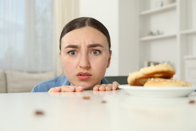 Photo of Shocked young woman looking at cockroaches and pretzels on white table indoors. Pest problem