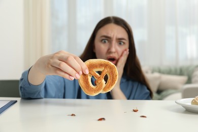 Photo of Shocked young woman holding pretzel with cockroach at white table indoors, selective focus. Pest problem