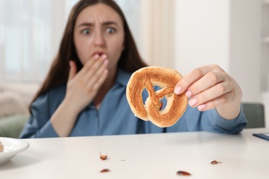 Photo of Shocked young woman holding pretzel with cockroach at white table indoors, selective focus. Pest problem
