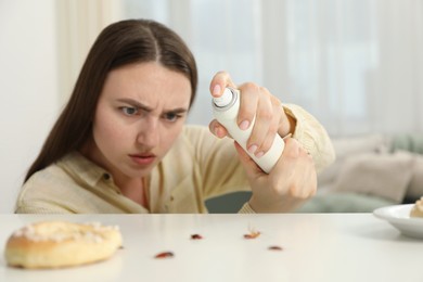 Photo of Emotional young woman spraying insecticide onto cockroaches at white table indoors, selective focus. Pest problem