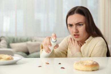 Photo of Emotional young woman spraying insecticide onto cockroaches at white table indoors, selective focus. Pest problem
